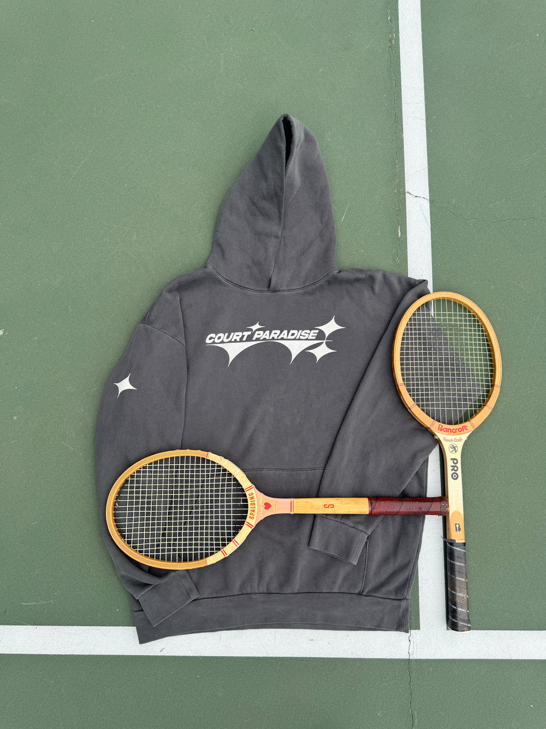 Court Paradise Charcoal hoodie laid flat on the court with two vintage wood tennis racquets, gift-worthy tennis apparel