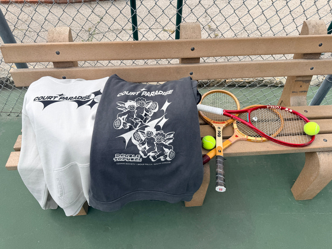 Court Paradise Bone and Charcoal hoodies draped on a public tennis court bench with vintage wood racquets, tennis streetwear