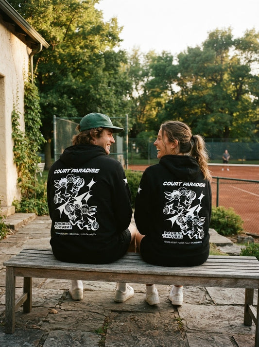 Two friends in black Court Paradise Double Trouble hoodies sit on a bench courtside, cherub tennis graphic on the back, clay courts behind them