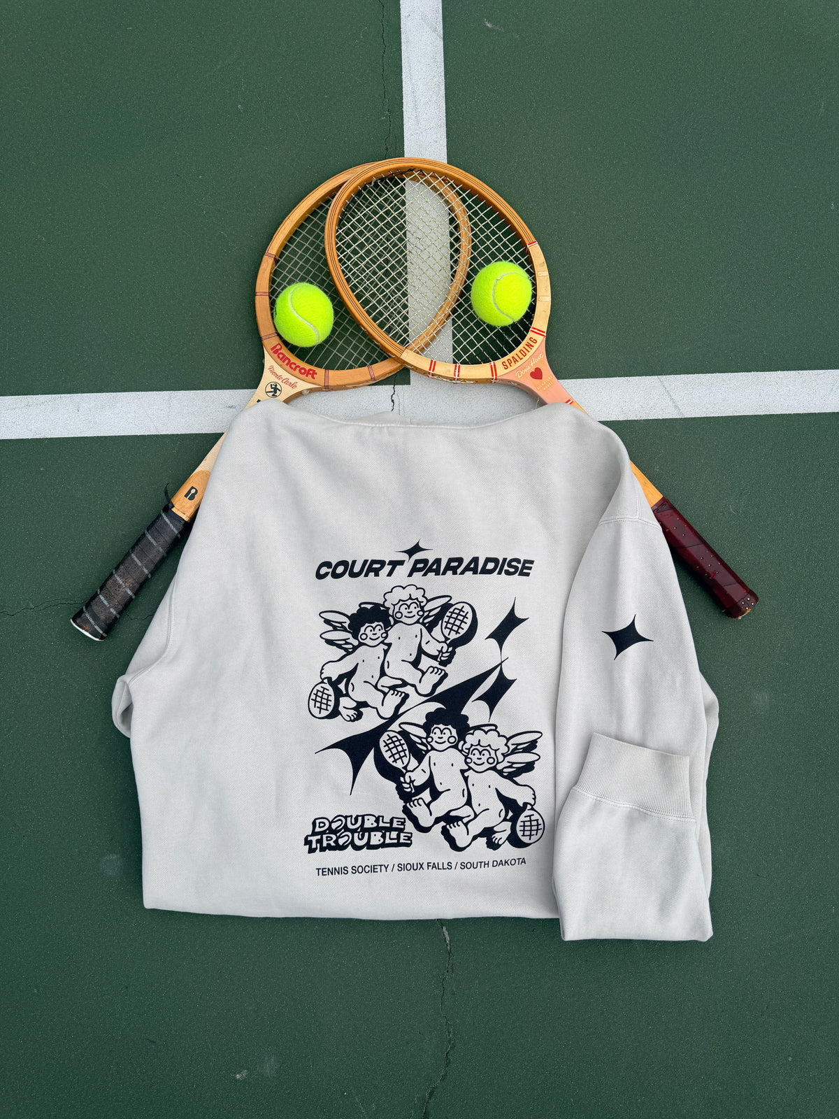 Double Trouble Hoodie in Bone laid on a green hard court, cherub back print, with wooden racquets and tennis balls