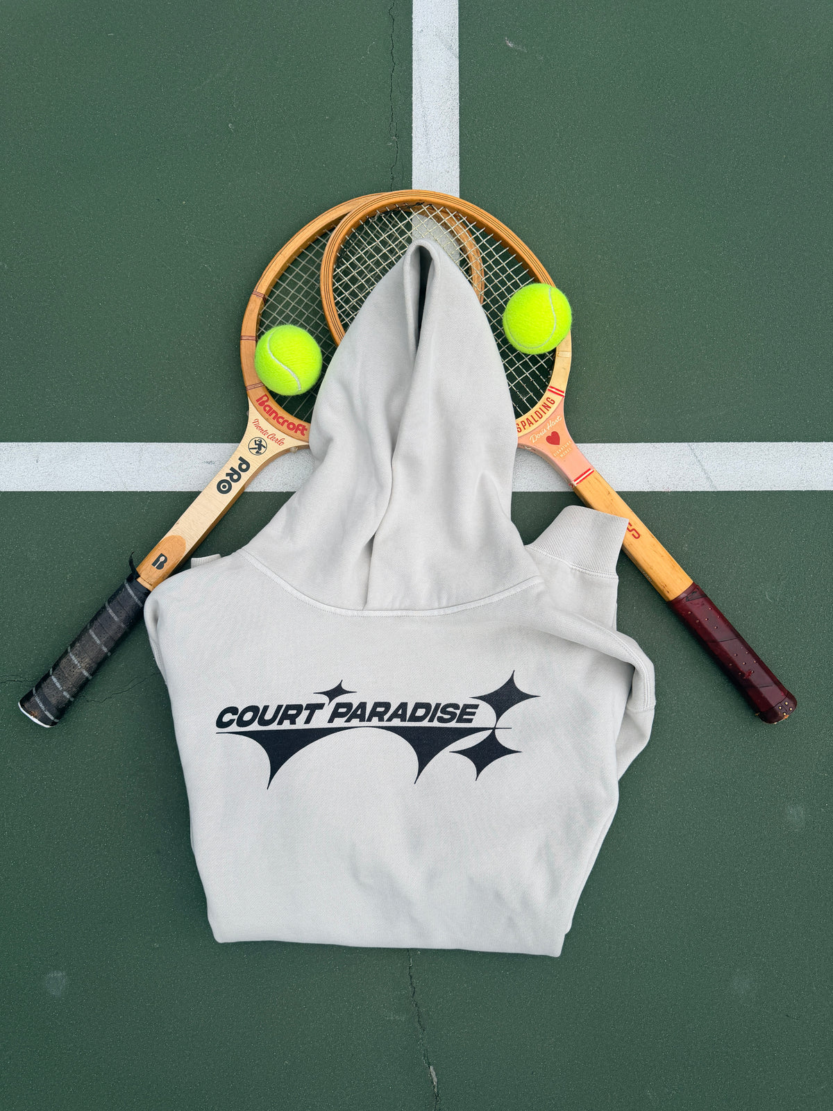 Double Trouble Hoodie in Bone on a green hard court, front chest wordmark, with wooden racquets
