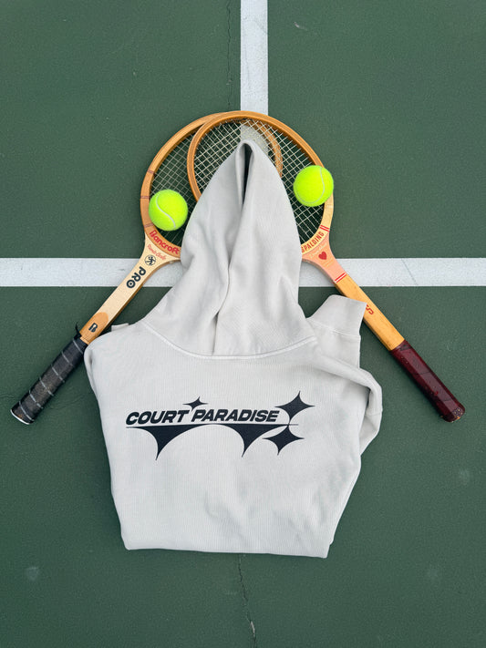 Double Trouble Hoodie in Bone on a green hard court, front chest wordmark, with wooden racquets