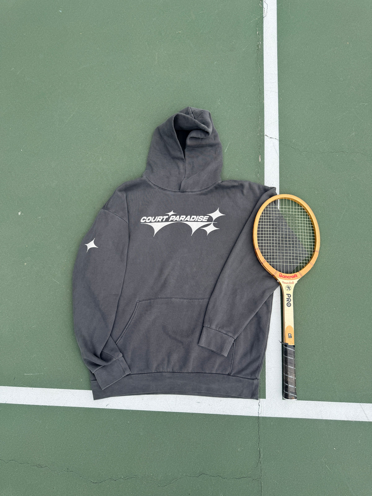 Double Trouble Hoodie in Charcoal on a green hard court, front chest wordmark, next to a wooden racquet
